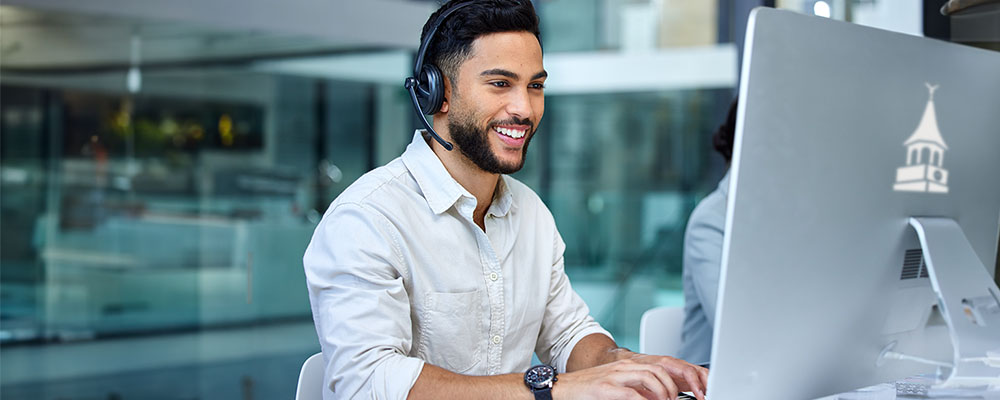Male counselor wearing headset, smiling while typing on an iMac.