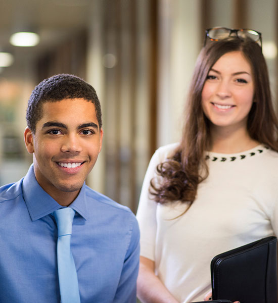 A man and woman student standing and smiling while wearing business attire.